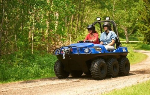 Two people ride in a blue all-terrain vehicle on a dirt path surrounded by greenery. Both are wearing helmets.