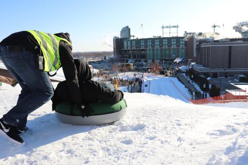 A person in a green tube is being pushed down a snowy slope, with a stadium and crowd visible in the background.