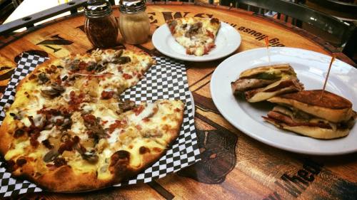 A table with a pizza topped with mushrooms and bacon, and two sandwiches on a plate, with condiments in the background.