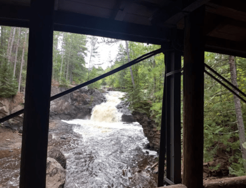 View of a waterfall cascading through a forest, framed by wooden beams and greenery.