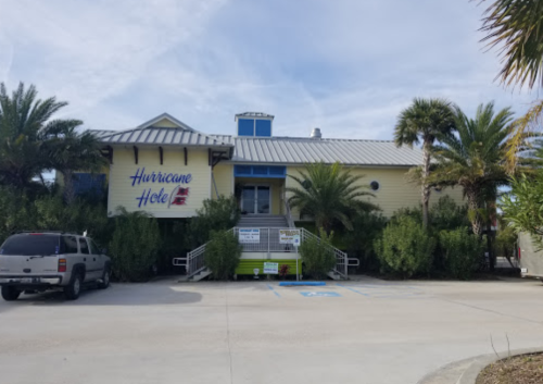 A colorful building named "Hurricane Hole" surrounded by palm trees and greenery, with a parking area in front.
