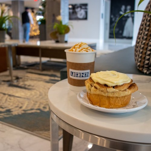 A cozy café scene featuring a cinnamon roll with frosting and a coffee cup topped with caramel drizzle on a table.