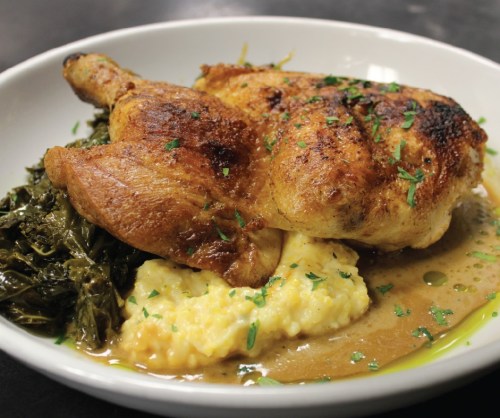 A plate featuring roasted chicken, creamy grits, and sautéed greens, garnished with fresh herbs.
