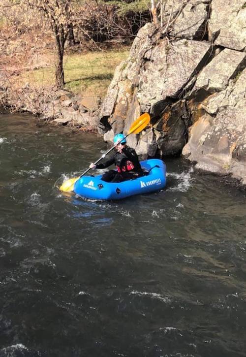 A person in a blue inflatable kayak paddles on a river near rocky banks, surrounded by trees.