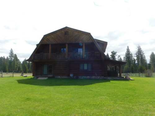 A large log cabin with a balcony, surrounded by green grass and trees under a cloudy sky.