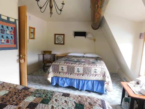 Cozy bedroom with a quilted bedspread, wooden chair, and a small TV, featuring sloped ceilings and natural light.