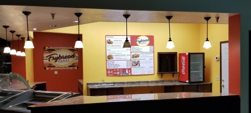 Interior of a restaurant with a colorful wall, menu board, and a soda fridge, featuring pendant lighting.