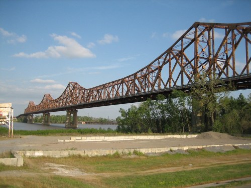 A rusted metal bridge spans a river, surrounded by green trees and a grassy area under a blue sky with clouds.