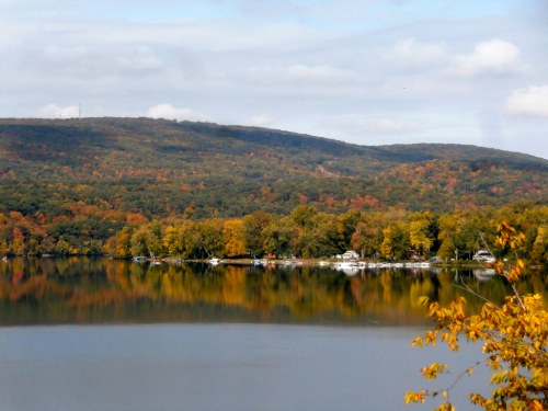 A serene lake reflects colorful autumn trees and hills under a partly cloudy sky. Boats are docked along the shore.
