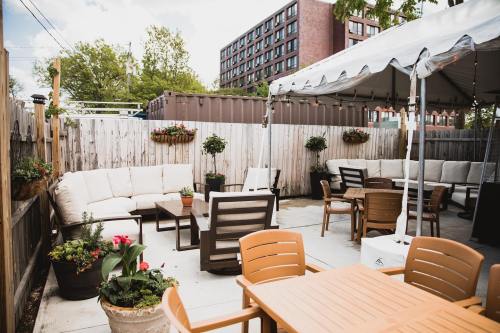 Outdoor patio with seating, tables, and potted plants, under a tent with string lights and a wooden fence in the background.