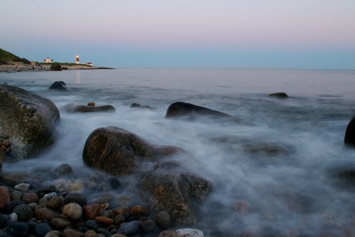 A serene coastal scene with smooth rocks in the foreground and a distant lighthouse against a pastel sky.