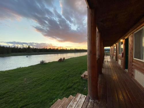 A wooden porch overlooks a river at sunset, with lush green grass and trees in the background.