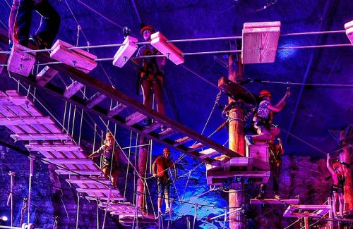 A group of people navigating a high ropes course with wooden platforms and safety harnesses in a dimly lit environment.