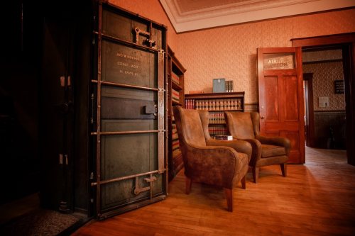A vintage room featuring two armchairs, a large safe, and wooden bookshelves in warm lighting.