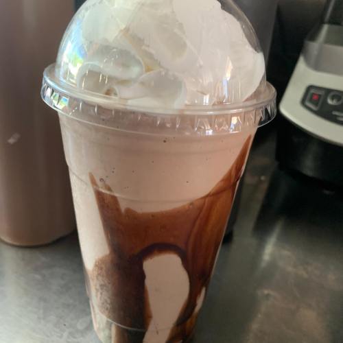 A clear cup filled with a creamy beverage, topped with whipped cream and drizzled with chocolate syrup.