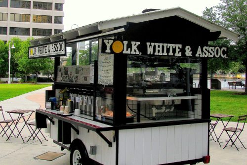 A food cart named "Yolk, White & Assoc" in a park, featuring a glass display and outdoor seating.