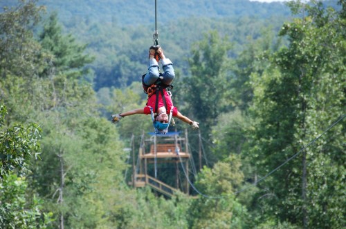 A person zip-lining through a forest, arms outstretched, with trees and a platform visible in the background.