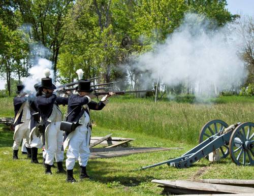 Historical reenactors in period uniforms firing muskets beside a cannon, with smoke and greenery in the background.