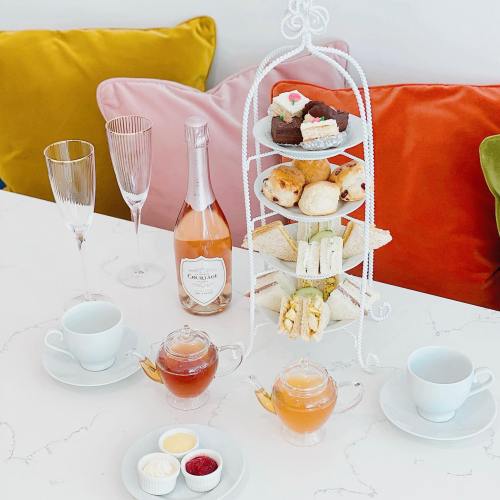 A tiered stand with pastries and sandwiches, two teapots, champagne flutes, and colorful cushions in the background.