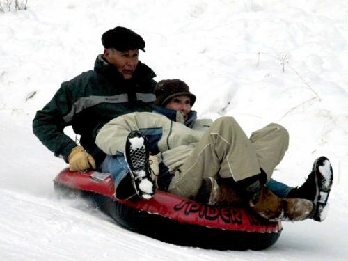 Two people joyfully tubing down a snowy hill, bundled in winter clothing, with snow-covered ground in the background.