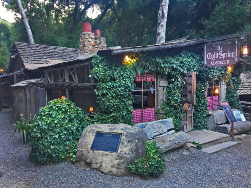 A rustic tavern covered in ivy, featuring a stone chimney and wooden sign, set in a wooded area.