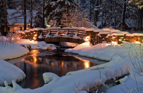 A serene winter scene featuring a snow-covered bridge over a calm river, illuminated by soft lights amidst trees.