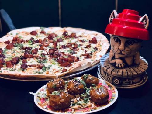 A delicious pizza topped with meat and herbs, alongside a plate of round snacks and a decorative cat figurine.