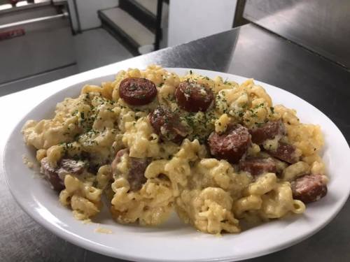 A plate of creamy macaroni and cheese with sliced sausage, garnished with parsley, on a stainless steel countertop.