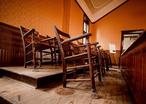 A row of vintage wooden chairs on a wooden floor, set against an orange wall with decorative patterns.