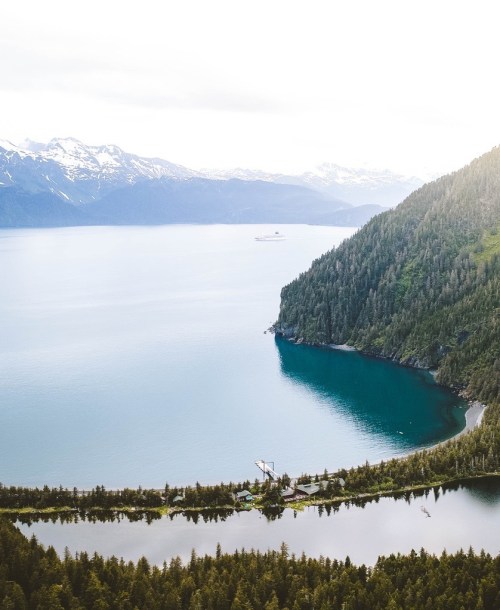 Aerial view of a serene lake surrounded by mountains and lush greenery, with a small dock and boat in the distance.