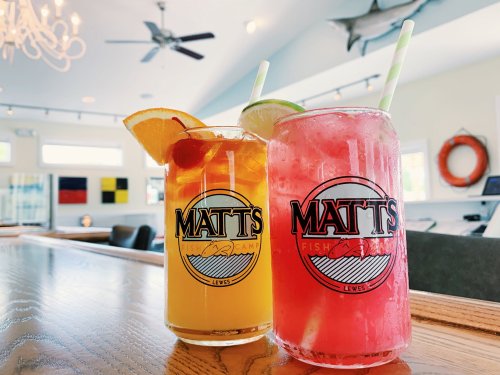 Two colorful cocktails with fruit garnishes sit on a wooden bar at Matt's Fish Camp, with a bright, airy background.