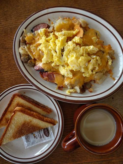 A plate of cheesy scrambled eggs, sausage, and potatoes, with toast and a cup of coffee on the side.