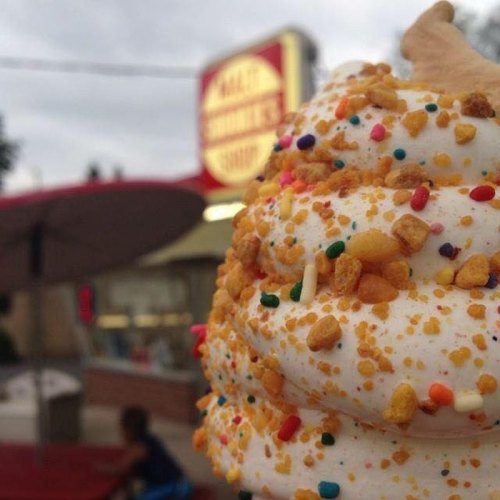 A close-up of a soft-serve ice cream cone topped with colorful sprinkles, with a shop in the background.