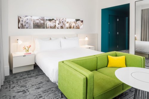 A modern hotel room featuring a white bed, green couch, and artwork of sheep on the wall. Bright and inviting decor.