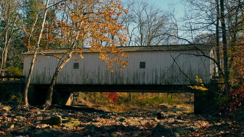 A weathered covered bridge spans a dry riverbed, surrounded by autumn foliage and bare trees.