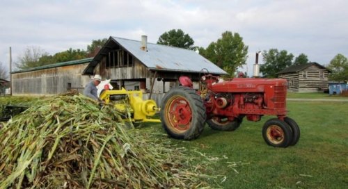 A farmer operates a yellow machine next to a red tractor, with a rustic barn in the background and green fields around.