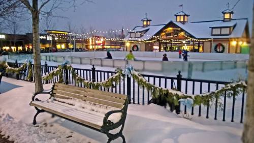 A snowy scene featuring a skating rink, decorated buildings, and a bench adorned with garlands and blue bows.