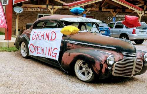 A vintage car with a "Grand Opening" sign and colorful balloons parked outside a community hall.