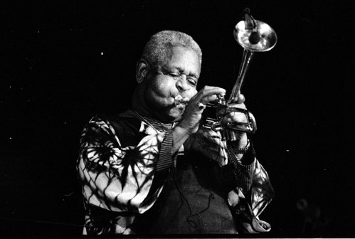 A musician passionately playing the trumpet on stage, wearing a patterned jacket, captured in black and white.