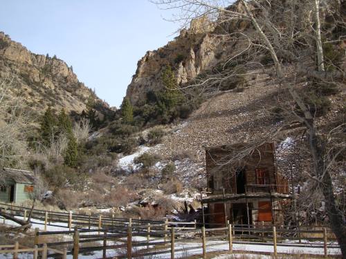 Abandoned wooden building near a rocky hillside, surrounded by trees and a wooden fence in a winter landscape.