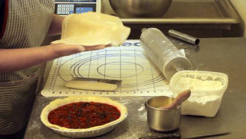 A person holds a pie crust above a filled pie dish, with flour and rolling pin on a countertop.