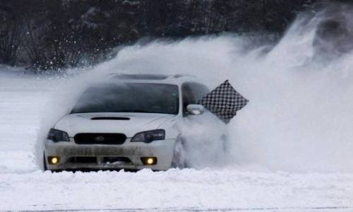 A white car drifts through deep snow, with a checkered flag waving from the window. Snow sprays around it.