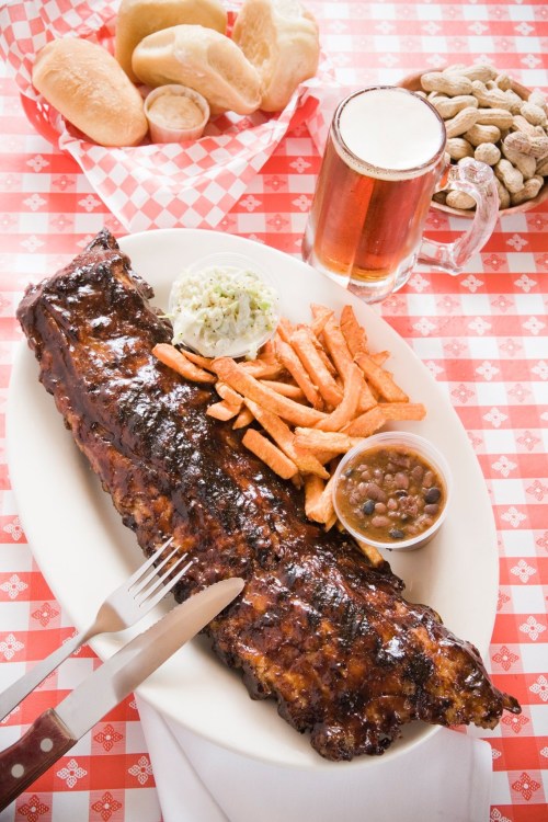 A plate of BBQ ribs with sweet potato fries, coleslaw, baked beans, rolls, and a mug of beer on a checkered tablecloth.