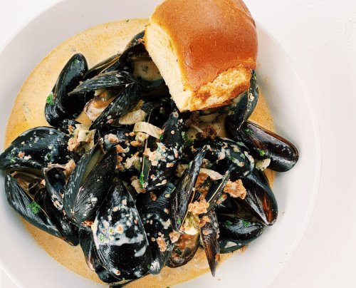 A bowl of mussels in a creamy sauce, topped with herbs, accompanied by a soft bread roll.