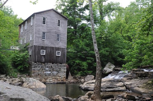 A historic wooden mill stands by a creek, surrounded by lush greenery and rocky terrain. Water flows nearby.