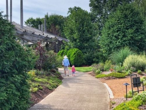 A person and a child walk hand in hand along a garden path surrounded by lush greenery and colorful plants.