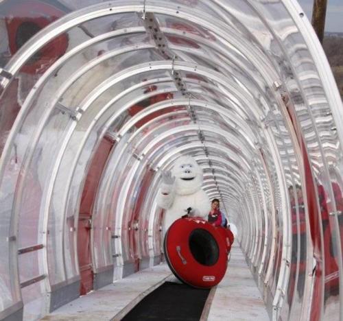 A yeti figure waves while sitting on a red inner tube in a clear, curved tunnel.