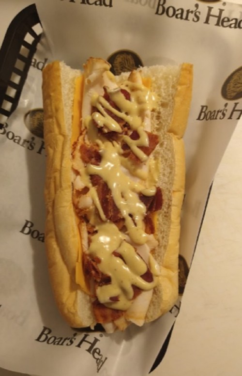 A sub sandwich with layers of turkey, bacon, cheese, and drizzled with mustard, served in a paper wrapper.