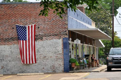 A brick building with a large American flag hanging, featuring a porch with chairs and a person sitting outside.