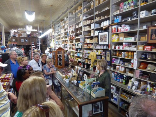 A crowd gathers around a woman speaking at a vintage store filled with various products and memorabilia on shelves.